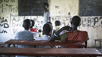 Auf einer hölzernen Schulbank sitzen in einem dunklen Klassenzimmer drei Jungen in T-Shirts nebeneinander und blicken zur Tafel und dem Lehrer