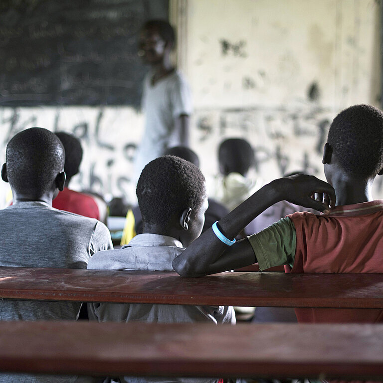 Auf einer hölzernen Schulbank sitzen in einem dunklen Klassenzimmer drei Jungen in T-Shirts nebeneinander und blicken zur Tafel und dem Lehrer