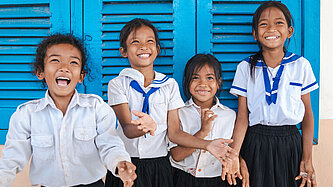 Vier lächelnde Mädchen in Schuluniform spielen vor einer blauen Wand mit einem Luftballon in Kambodscha.