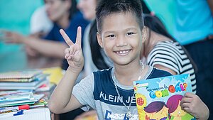 Bildung in Vietnam: Schulkind mit Heft Lachender Junge zeigt Peace-Zeichen und hält ein Heft im Klassenzimmer in Vietnam.
