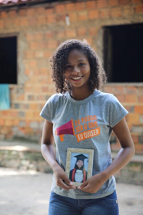 Eine junge Frau steht im T-Shirt vor einem Gebäude, sie hält ein Bild von sich beim Schulabschluss in der Hand