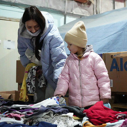 Ein Mädchen und ihre Mutter sind in einem Lagerraum und schauen sich Anziesachen an, die vor ihnen ausgelegt sind.