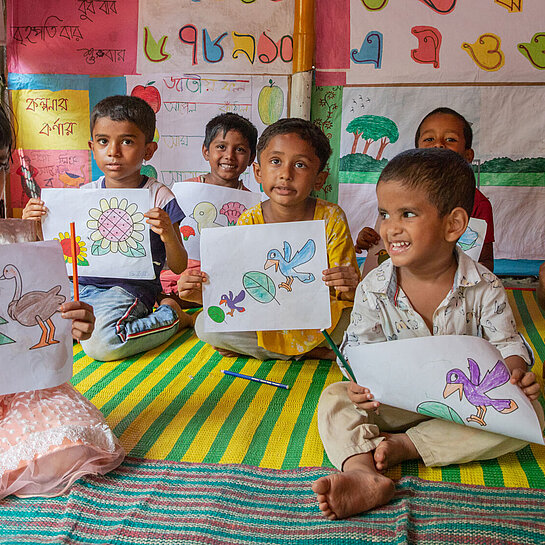 Sieben kleine Kinder sitzen auf Teppichen auf dem Boden und halten bunt bemalte Bilder in den Händen.
