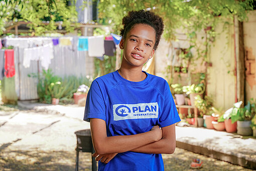 Ein Mädchen trägt ein Plan-T-Shirt, hat die Arme vor der Brust verschränkt und schaut in die Kamera