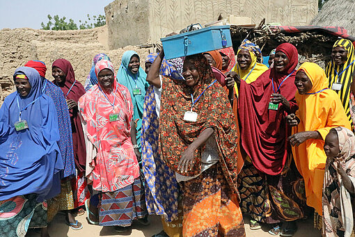 Einmal pro Woche treffen sich die Frauen der Spargruppe Akula da Kyau – und investieren in die Zukunft ihrer Töchter Eine Gruppe von Frauen in bunten Gewändern hat sich auf einem Dorfplatz versammelt. Eine Frau in der Mitte der Gruppe trägt eine metallene Geldkassette auf dem Kopf.