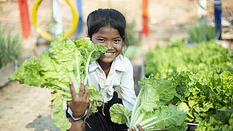 Ein fröhliches Mädchen aus Kambodscha hält zwei frische Kopfsalate in einem Gemüsegarten und lächelt.