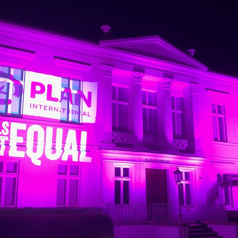 Pink beleuchtetes Gebäude mit projizierter Schrift darauf (Plan Logo und Girls get equal)