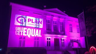 Pink beleuchtetes Gebäude mit projizierter Schrift darauf (Plan Logo und Girls get equal)