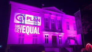 Pink beleuchtetes Gebäude mit projizierter Schrift darauf (Plan Logo und Girls get equal)