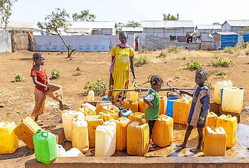 Mehrere Personen stehen an einer Anlage zur Wasserversorgung und befüllen gelbe Kanister mit Wasser