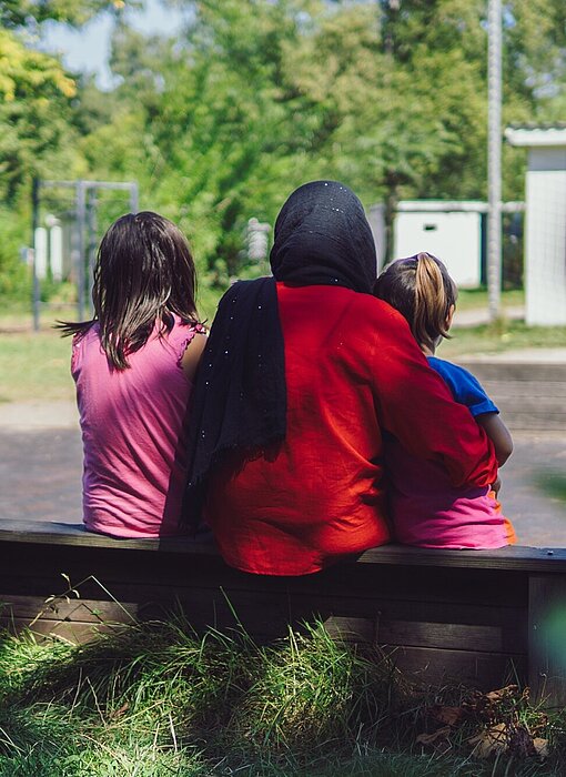 Eine Frau sitzt mit zwei Kindern im Arm auf einer Mauer, von hinten fotografiert