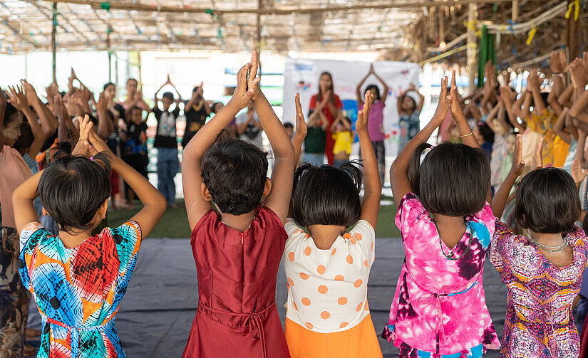 Mehrere Kinder, überwiegend Mädchen, stehen in bunter Kleidung mit dem Rücken zur Kamera in einer großen, offenen Halle. Sie haben die Arme über den Kopf erhoben und die Hände zum Applaus zusammengelegt, als Teil einer gemeinsamen Aktivität, vermutlich Tanz oder Bewegungsspiel. Auch viele weitere Kinder und Erwachsene im Hintergrund machen die gleiche Geste.