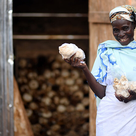 Eine Frau steht vor einem kleinen Haus. Sie hält Plastiktüten in der Hand, die voll mit kleinen, hellen Pilzen sind.