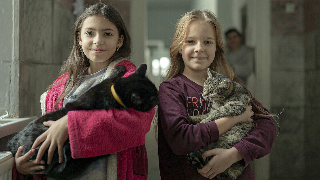 Zwei Mädchen stehen in einem Flur und lächeln in die Kamera. Sie haben zwei Katzen im Arm