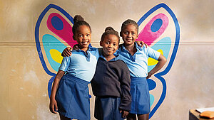 Mädchen weltweit sollen sich frei entfalten können - wie ein Schmetterling Mädchen weltweit sollen sich frei entfalten können. Drei Mädchen vor einer bunten Wand mit Schmetterlingsflügeln in einer Schule, die mit einem Lächeln posieren