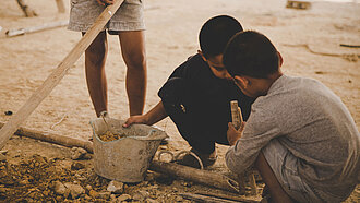 zwei Kinder sitzen mit einem kaputten Eimer auf sandigem Boden