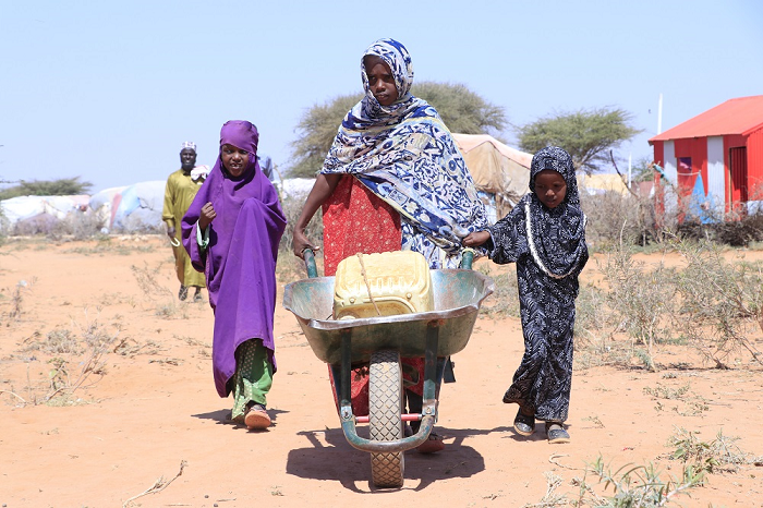 Eine Frau und zwei Mädchen transportieren einen Wasserkanister in einer Schubkarre