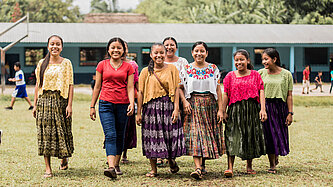 Eine Gruppe fröhlicher Mädchen in traditioneller Kleidung und modernen Outfits läuft über einen Schulhof in Guatemala