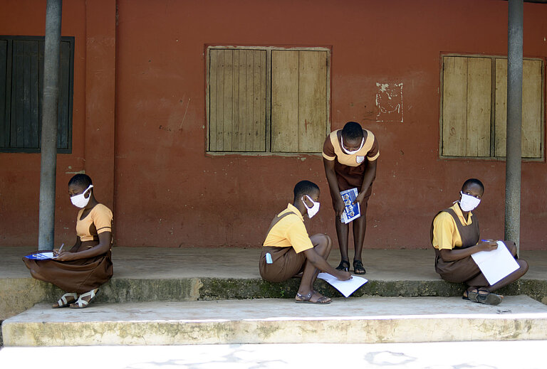 Vier Mädchen in Schuluniform sitzen vor einer Schule und lösen Aufgaben in Büchern.