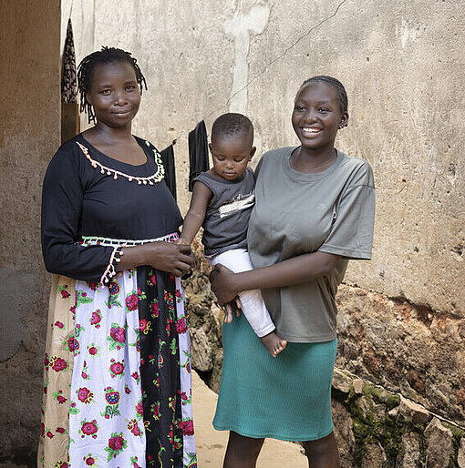 Faith mit ihrer Tochter im Arm und ihrer Mutter in ihrem Zuhause stehend