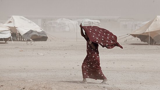 Eine junge Frau aus Burkina Faso läuft durch starken Wind, hält dabei ein Tuch über den Kopf