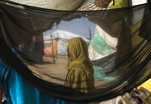 Eine sudanesische Frau steht in einem Geflüchtetencamp, ihr Gesicht ist nicht zu erkenne. Sie ist durch ein Zelt hindurch fotografiert.