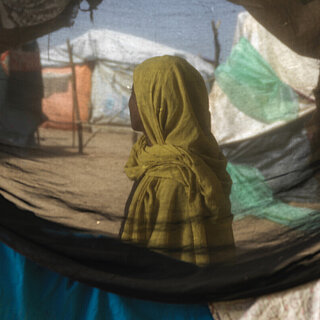 Eine sudanesische Frau steht in einem Geflüchtetencamp, ihr Gesicht ist nicht zu erkenne. Sie ist durch ein Zelt hindurch fotografiert.