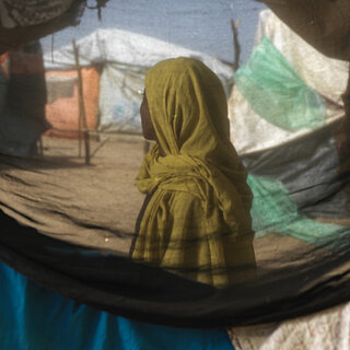 Eine sudanesische Frau steht in einem Geflüchtetencamp, ihr Gesicht ist nicht zu erkenne. Sie ist durch ein Zelt hindurch fotografiert.
