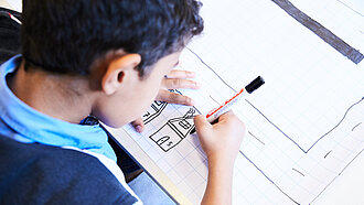 Ein Kind malt ein schwarzes Haus auf ein Blatt Papier.