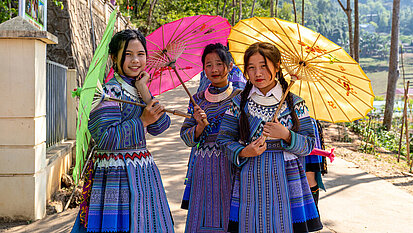 Drei jugendliche Mädchen aus Vietnam tragen traditionelle Kleidung und je einen bunten, traditionellen Sonnenschirm.