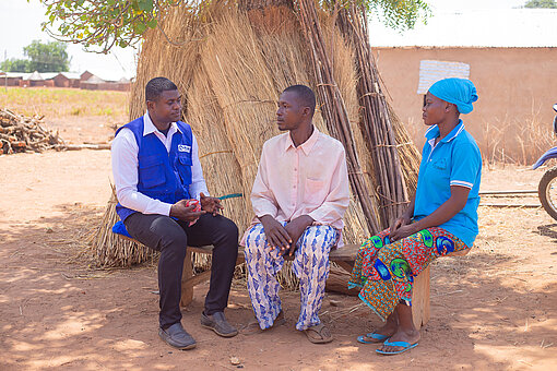 Drei ghanaische Menschen sitzen auf einer Holzbank. Eine Person trägt ein Kopftuch, eine andere eine blaue Weste von Plan Interational. Die drei sind im Gespräch