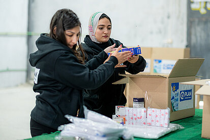 Nothilfe im Kontext des Nahost-Konflikts: Verpackung von Hilfspaketen Zwei Freiwillige verpacken Hilfspakete in einem Lagerraum im Kontext des Nahost-Konflikts. Sie halten Produkte in den Händen und bereiten die Pakete für die Verteilung vor.