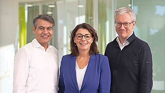 Zwei Männer, eine Frau stehen beisammen und schauen in die Kamera