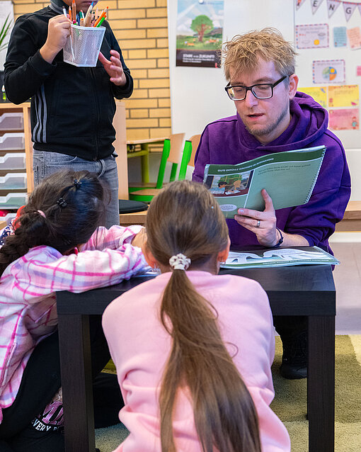 Workshopleiter Philipp Wolf liest gemeinsam mit zwei Schülerinnen im Rahmen des Projekts "Stark in die Zukunft" zur Förderung der mentalen Gesundheit von Grundschulkindern.