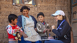 Mitarbeiterin einer Hilfsorganisation spricht mit Mutter und zwei Kindern vor einem Haus in einer ländlichen Gemeinde in Bolivien