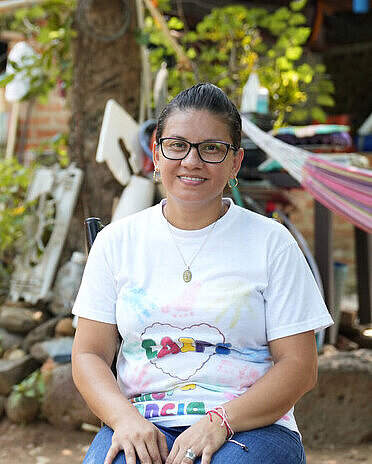 Mit ihrer Arbeit als Frühpädagogin stärkt Nancy Familien in Chalatenango. Eine Frau sitzt in einem Garten und lächelt in die Kamera. Hinter ihr ist eine bunte Hängematte angebracht.
