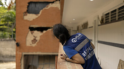 Eine Person mit blauer Weste von „Plan International“ und der Aufschrift „Response Humanitarian“ lehnt an einem Geländer und blickt auf ein stark beschädigtes Gebäude mit großen Löchern und abgebrochener Fassade.