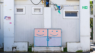 Ein Containergebäude mit einer bemalten Versorgungsbox, auf der zwei lächelnde Gesichter zu sehen sind.