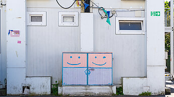 Ein Containergebäude mit einer bemalten Versorgungsbox, auf der zwei lächelnde Gesichter zu sehen sind.