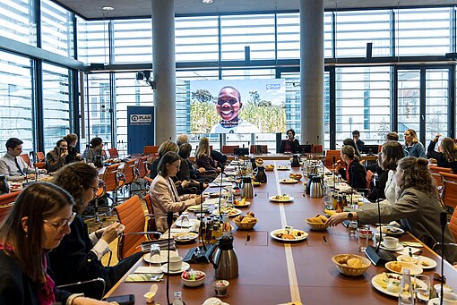 Blick über den langen Tisch des Parlamentarischen Frühstücks, am Ende der langen Tischreihe ist ein großer Bildschirm, auf dem ein Film mit Mädchen aus Simbabwe gezeigt wird