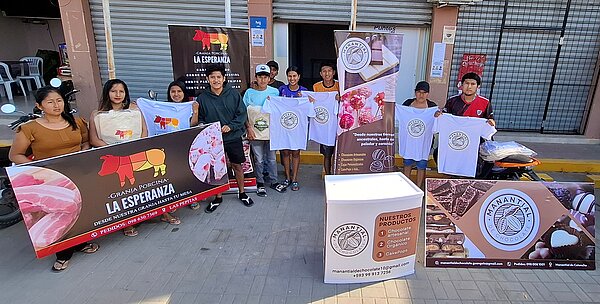 Eine Gruppe von Menschen steht vor einem Geschäft oder Verkaufsstand. Sie präsentieren Produkte wie T-Shirts und Werbebanner, die zu einer Marke gehören. Im Hintergrund sind Motorräder und geschlossene Läden sichtbar.