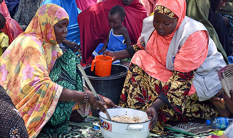 Zwei Frauen sitzen vor einem Topf und kochen, im Hintergrund sitzen weitere Frauen im Schatten eines Baumes