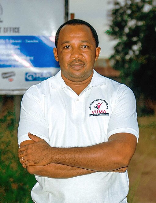 Ein Mann im weißen Polo-Shirt steht mit verschränkten Armen vor einer Tafel mit dem Logo von Plan International