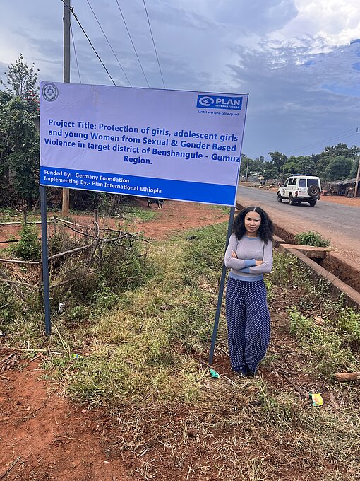 Jane Zentgraf steht vor einem Schild zu unsere Projekt in der Region Benshangule-Gumuz, Äthiopien