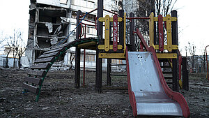 Ein Spielplatz vor einem zerstörten Gebäude im Kontext des Ukraine-Kriegs. Der Spielplatz ist leer, und die Zerstörungen im Hintergrund sind deutlich sichtbar.