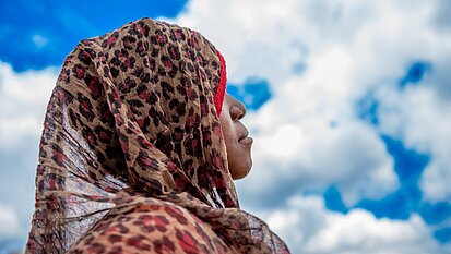 Ein 14-jähriges Mädchen aus Mosambik guckt in den strahlend-blauen Himmel mit weißen Wolken. Sie trägt ein Kopftuch mit Leopardenmuster, das ihr Haar bedeckt. Ihr Gesicht ist nicht zu sehen.