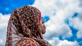 Ein 14-jähriges Mädchen aus Mosambik guckt in den strahlend-blauen Himmel mit weißen Wolken. Sie trägt ein Kopftuch mit Leopardenmuster, das ihr Haar bedeckt. Ihr Gesicht ist nicht zu sehen.