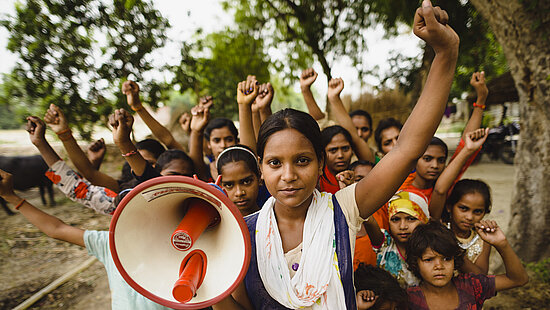 Eine Gruppe indischer Mädchen steht in einer Gruppe zusammen, sie haben jeweils eine Faust in die Höhe gestreckt. Das vorderste Mädchen hält ein Megaphon in der Hand.