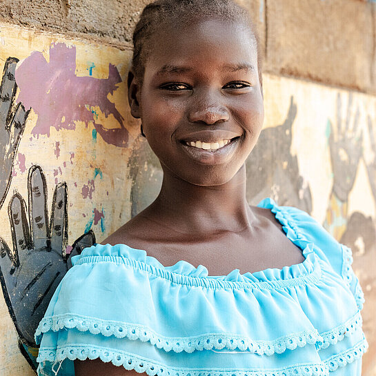 Lächelndes Mädchen in hellblauem Kleid steht mit verschränkten Armen vor einer bunt bemalten Wand mit Handabdrücken.
