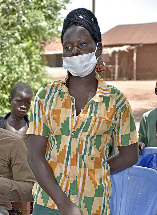 Ava, eine junge Frau, steht vor einer Gruppe Menschen und spricht. Sie trägt eine Mund-Nasen-Bedeckung.